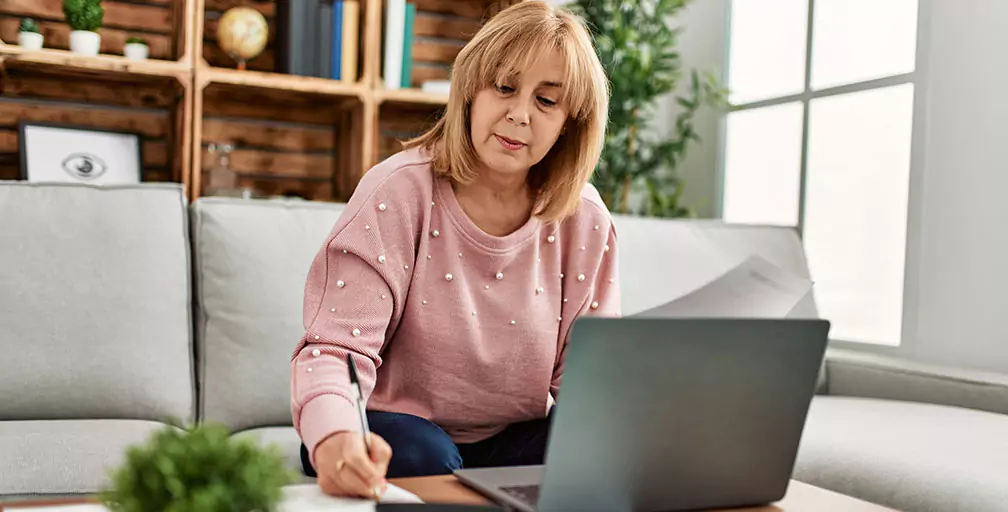 Sitting on her couch in front of a laptop on the coffee table, a woman jots down a note on a piece of paper. Experienced lawyers can help you apply for disability in Georgia.