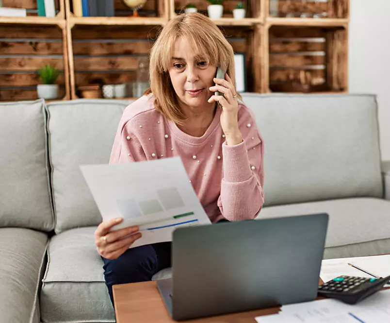 A woman talks on the phone and looks at a sheet of paper as she sits on a couch with her laptop in front of her. Disability applications require detailed information.