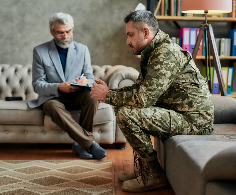 While a therapist listens, a solider in fatigues sits on the edge of couch, holding his hands together and looking downward. The VA attorneys at Silver & Archibald help with many aspects of the VA Disability system.