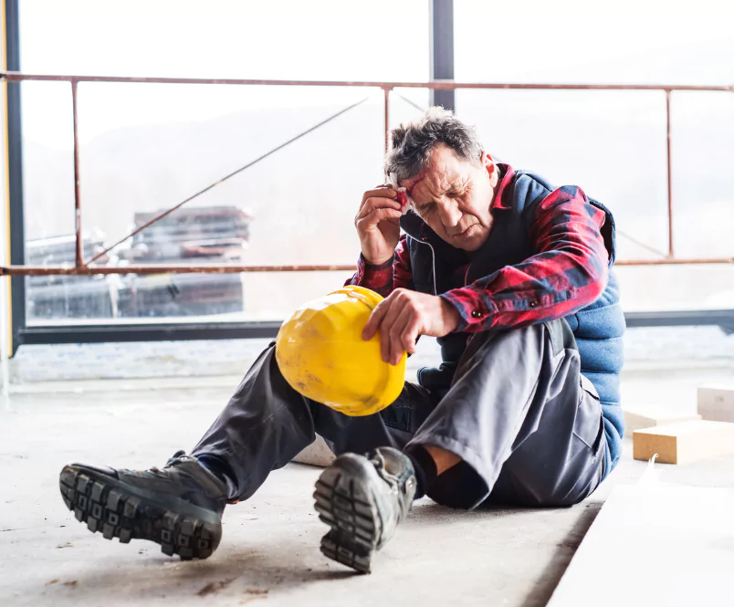 Sitting on the floor in a building under construction, man takes off his hardhat and dabs an injury on his forehead. For job injuries, workers’ compensation benefits provide medical and financial help.
