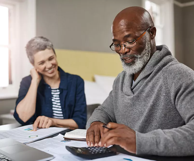 Smiling, a man taps on a calculator on a desk full of papers, while a woman, also smiling, looks on. You talk to Silver & Archibald lawyers about your workers’ comp or personal injury case